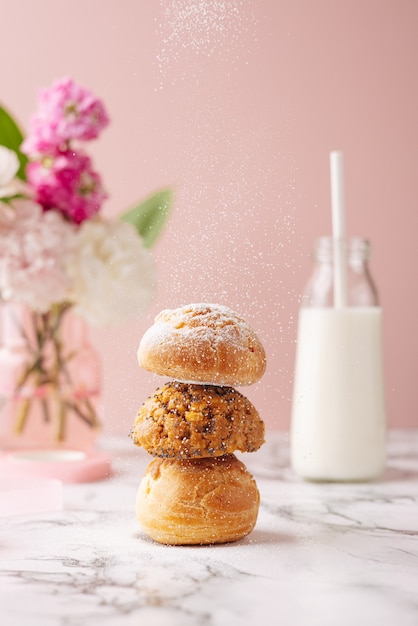 Homemade profiteroles with custard covered sugar powder on marble table on pink background with flowers and milk vertical