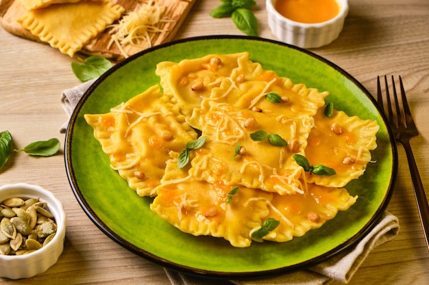 Homemade Pumpkin Ravioli with ricotta cheese served basil, pine nuts and hard cheese on green plate surrounded by ingredients on wooden table. Autumn food concept. Selective focus.