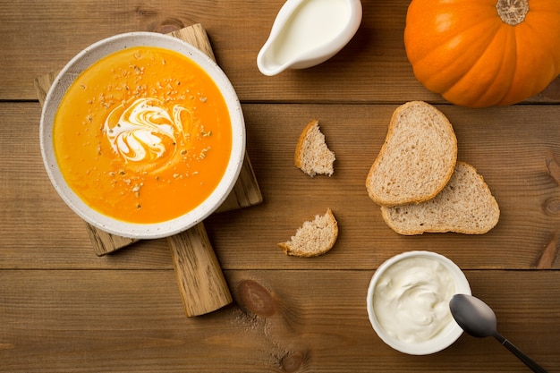 Homemade pumpkin soup in white plate with cream and bread on cutting board flat lay on wooden background