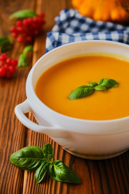 Homemade pumpkin soup with cream, croutons, pumpkin seeds and basil on rustic wooden background, selective focus