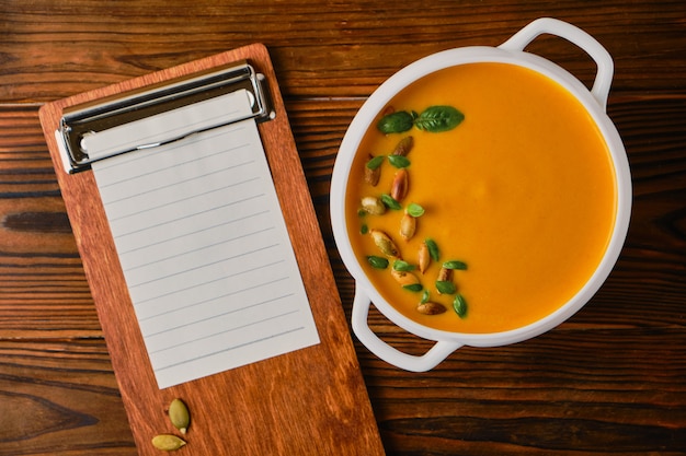 Homemade pumpkin soup with cream in a white bouillon dish with seeds and basil, Wooden tablet with a sheet of paper for a recipe, selective focus, wooden background