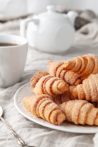 Homemade rollshaped shortbread Sprinkled with sugar On a white plate Background gray linen In the background is a white teapot and a cup Country style