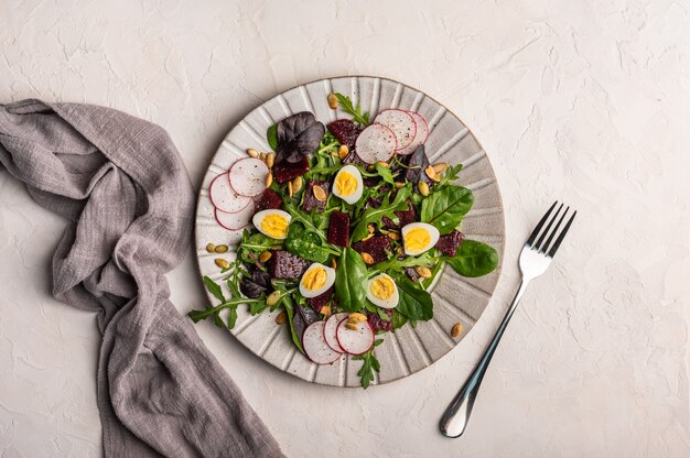 Homemade salad of quail eggs, boiled beetroot, radish, arugula and basil with olive oil served with linen napkin and fork on a light background, top view, copy space