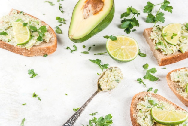 Homemade sandwich toasts with with guacamole, lime lemon and parsley on white table, copy space