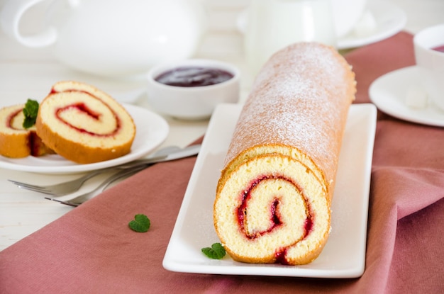 Homemade sponge cake roll with raspberry jam on a white plate with a cup of fragrant berry tea