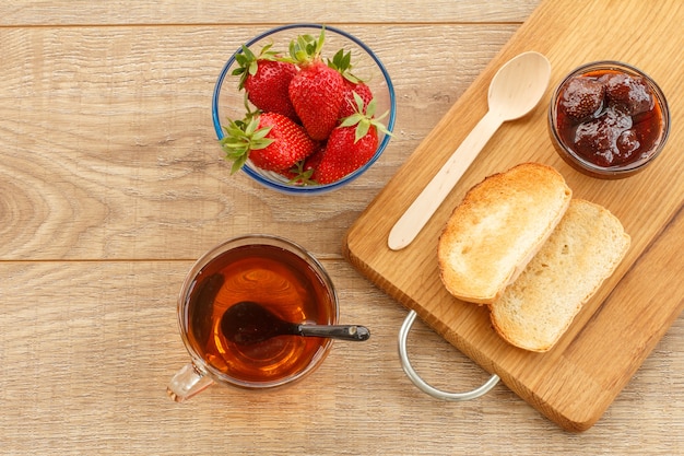 Homemade strawberry jam in a glass bowl and toasts on cutting board, fresh berries in bowl, cup of tea on the wooden background. Top view.