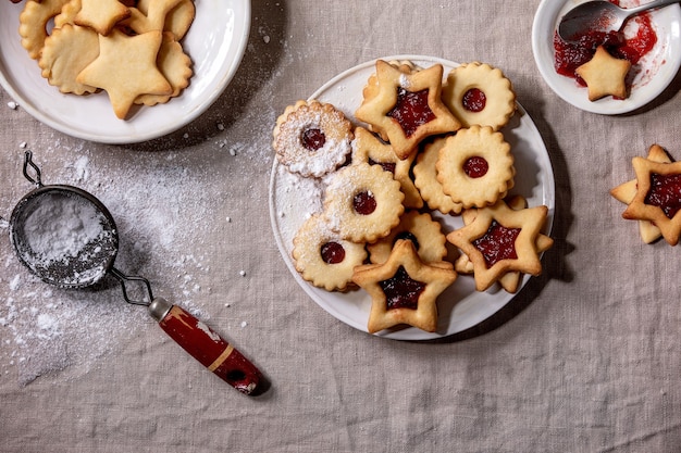 Homemade traditional Linz shortbread biscuits cookies with red jam and icing sugar on ceramic plate over linen tablecloth. Flat lay, space