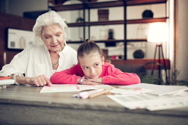 Homework together. Dark-eyed girl sitting near her loving caring granny while doing homework together