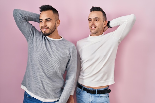 Homosexual couple standing over pink background smiling confident touching hair with hand up gesture, posing attractive and fashionable