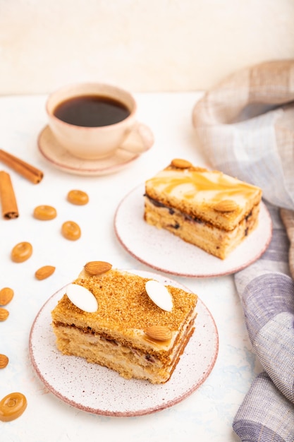 Honey cake with milk cream, caramel, almonds and a cup of coffee on a white concrete background and linen textile. Side view, selective focus, close up.