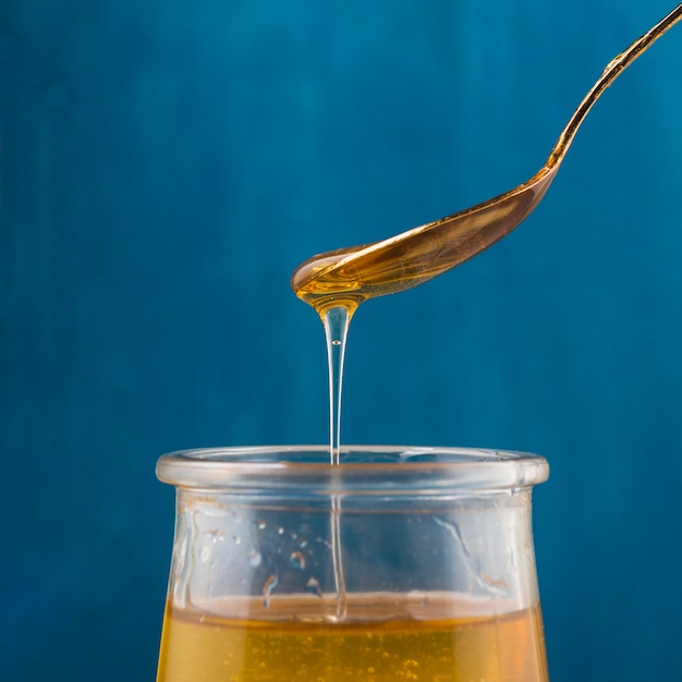 Honey dripping from spoon in glass pot against blue background