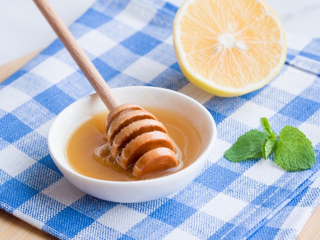 Honey in white bowl with honey dipper or stick, lemon and mint in blue and white linen towel