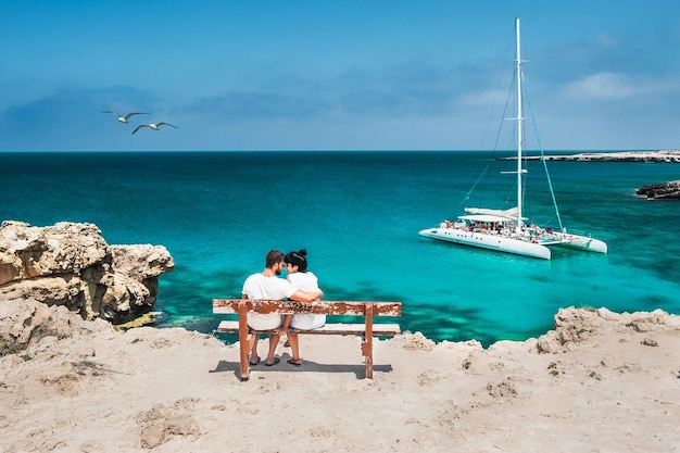 Honeymoon traveller couple hugging on a wooden bench and enjoys their tropical holiday. Wedding travel. Young happy couple sitting back on beach. Rear view of a couple in love on vacation. Travelers