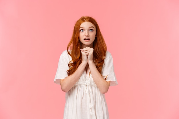 Hopeful and amused, redhead female in white dress, holding hands together over chest