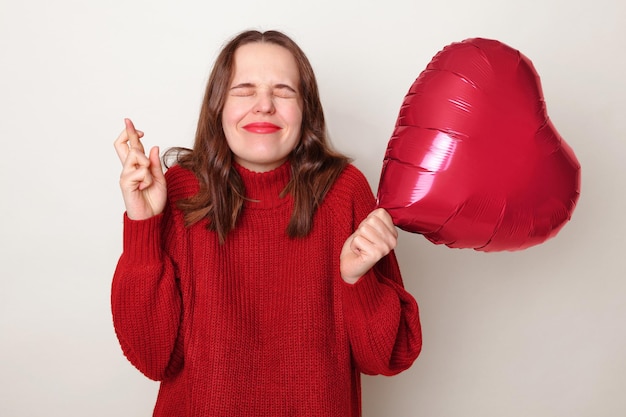 Hopeful Caucasian woman wearing red sweater holding heart shaped balloon isolated over gray background crossing finger making wish wants to receive invitation for dating on Valentines Day