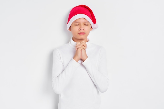 Hopeful little Asian man in christmas hat keeps palms pressed together in praying gesture isolated over white background Happy New Year 2023 holiday concept