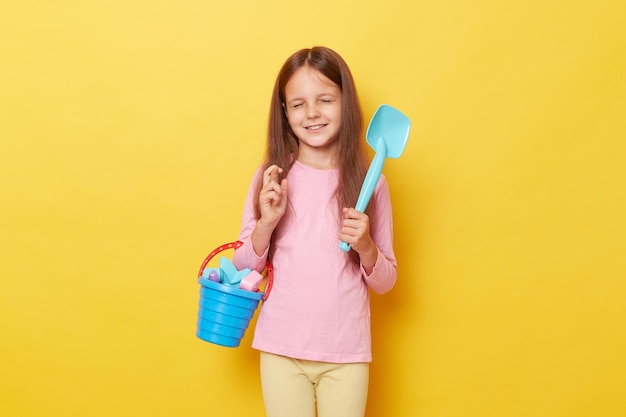 Hopeful little girl wearing casual clothing holding sandbox toys and bucket isolated over yellow background crossing fingers wants to go to play outdoor in sand with puddle