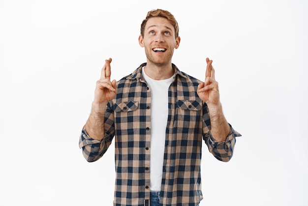 Hopeful redhead man looking up with crossed fingers praying or waiting for miracle making wish anticipating good news standing over white background