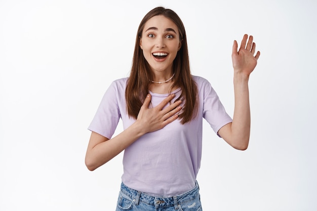 Hopeful smiling girl looks excited, raising hand and place one on chest, introduce herself, volunteer, wants to be picked or chosen, standing on white