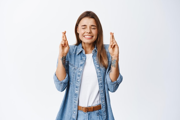 Hopeful young woman believe dream come true, cross fingers for good luck with closed eyes and excited face, standing on white