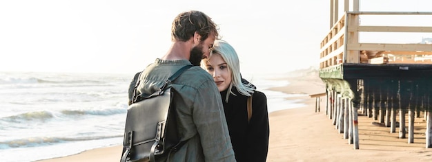 Horizontal banner or header with romantic hipster couple in love standing on shoreline at the wild beach near the wooden structure Boyfriend and girlfriend wearing cool clothing and vintage backpack