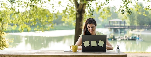 Horizontal banner with young woman using laptop computer in the public park Millennial female freelancer working remotely and taking notes in a city park Technology and remote work concept