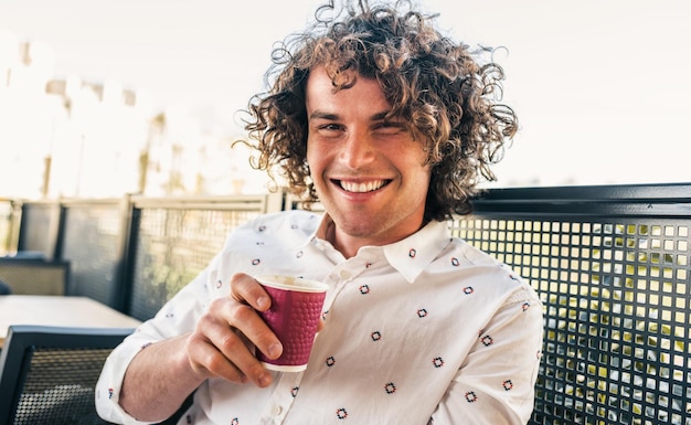 Horizontal closeup portrait of male with toothy smile curly hair wearing casual trendy shirt drinking a cup of coffee in the city cafe Businessman have a coffee break People and lifestyle concept