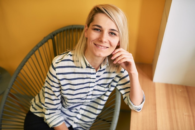 Photo horizontal closeup portrait of young beautiful blonde woman resting on the modern chair and looking to the camera in the office pretty business female wearing striped shirt sitting in workplace