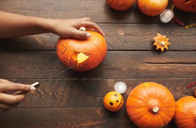 Horizontal from above view shot of hands carving ripe pumpkin for Hallowing party with kitchen knife on dark brown wooden table