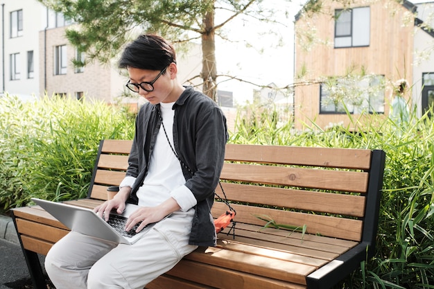 Horizontal medium portrait of modern young Asian woman wearing casual outfit with eyeglasses sitting on bench outdoors doing some freelance work using laptop