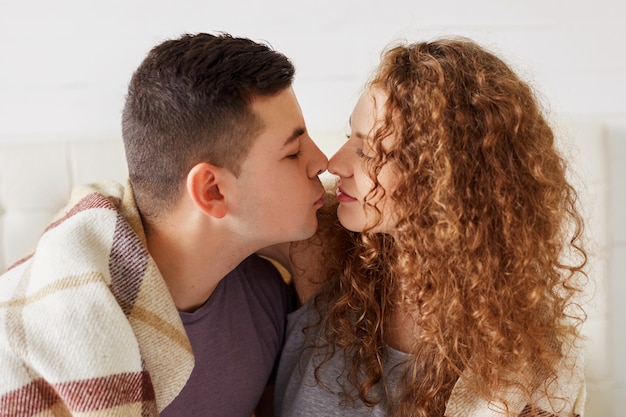 Horizontal portrait of affectionate newlywed couple touch noses and going to kiss, enjoy domestic calm atmosphere, covered with warm plaid