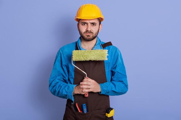 Horizontal portrait of displeased upset sad builder having frowned facial expression, having problems, holding roller in both hands. People and crisis concept.