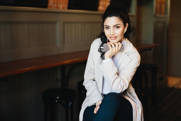 Horizontal portrait of fashionable woman with dark eyes and hair having pure skin looking in camera sitting in cafe enjoying good music while listening to her favorite tracks in white earphones