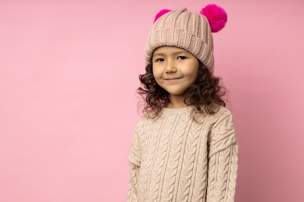 Horizontal portrait of friendly little girl with curly hair, wearing beige sweater, winter hat with crimson furry pom poms, smiling looking, with copy space. Kids fashion.