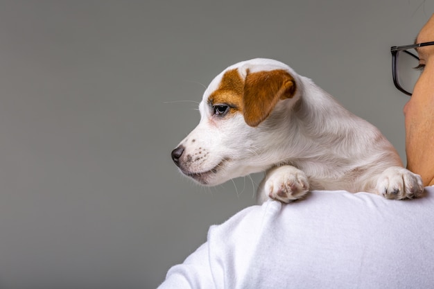 Horizontal portrait of handsome cheerful man holds jack russell terrirer on his shoulder, has glad expression