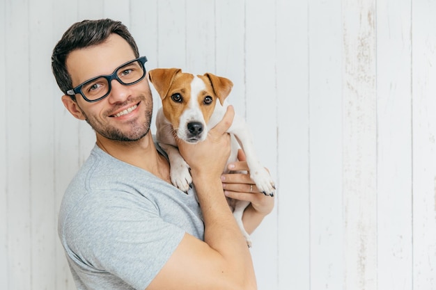 Horizontal portrait of handsome cheerful man wears eyeglasses holds jack russell terrirer has glad expression poses against white wooden wall with blank copy space Animals and friendship