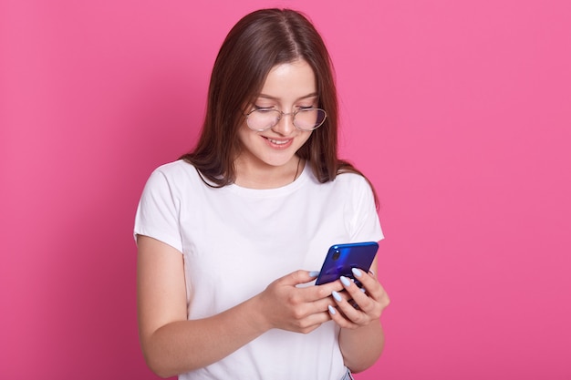 Horizontal shot of adorable teen with long hair, holding modern cell phone, scrolls through social networks, has cheerful expression, wears spectacles and casual t shirt, isolated over pink