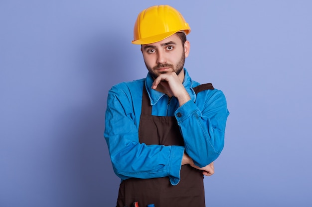 Horizontal shot of attentive serious young handyman having pensive facial expression, , wearing apron, helmet and overalls, having thoughts about project.