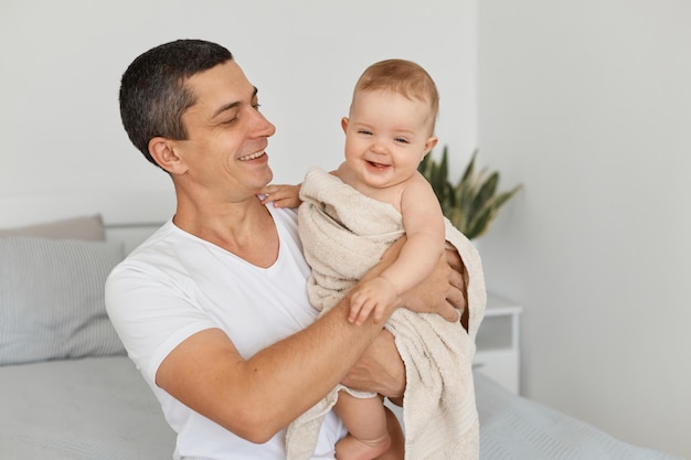 Horizontal shot of attractive brunette man wearing white casual t shirt holding his infant daughter, posing at home in light room, looking at his kid, expressing happiness.