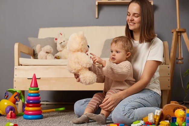 Horizontal shot of baby and mother sitting on floor at home ans playing with soft teddy bear family sitting together and expressing positive emotions posing surrounded with colorful toys