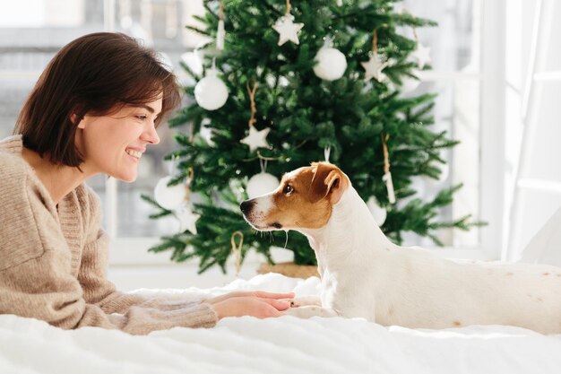 Horizontal shot of beautiful cheerful woman enjoys togetherness with dog holds its paws lie on white bed look each other in eyes feel love and friendship decorated Christmas tree in background