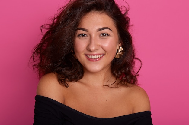 Horizontal shot of beautiful young woman, looking smiling at camera, posing with bared shoulders, dressed black shirt, has happy facial expression, adorable lady stands isolated over pink studio wall.