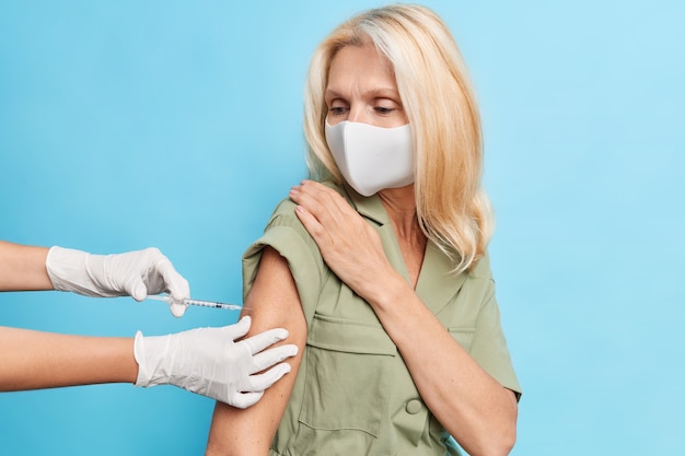 Horizontal shot of blonde forty years old woman wears disposable mask to protect herself from coronavirus gets injection in clinic isolated on blue wall