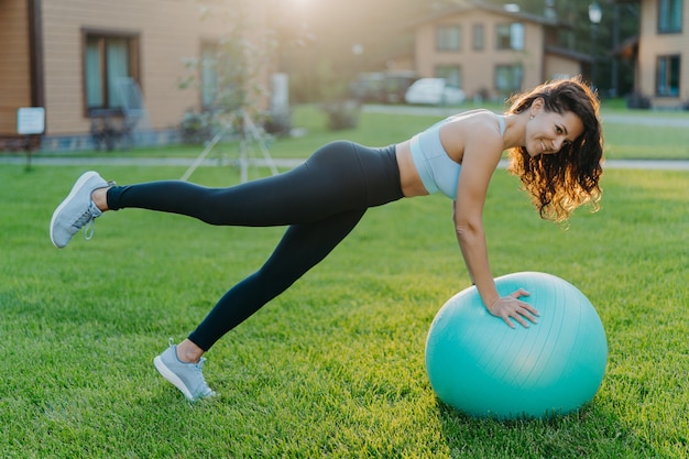 Horizontal shot of brunette woman leans on fitness ball, raises legs and does gymnastic exercises, wears top leggings and sneakers, poses in yard near house during good sunny day, breathes fresh air