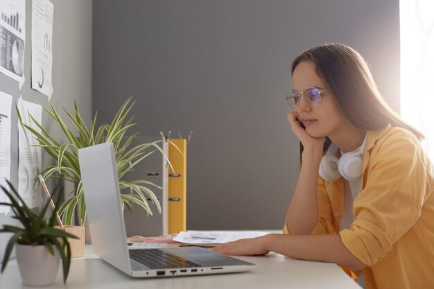 Horizontal shot of calm beautiful woman dressed in yellow shirt using laptop computer and sitting at table in office looking at notebook monitor learning information