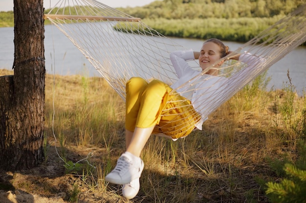 Horizontal shot of Caucasian happy delighted woman sitting on hammock by the water posing with raised arms and relaxing on the bank of the river camping having rest on weekend