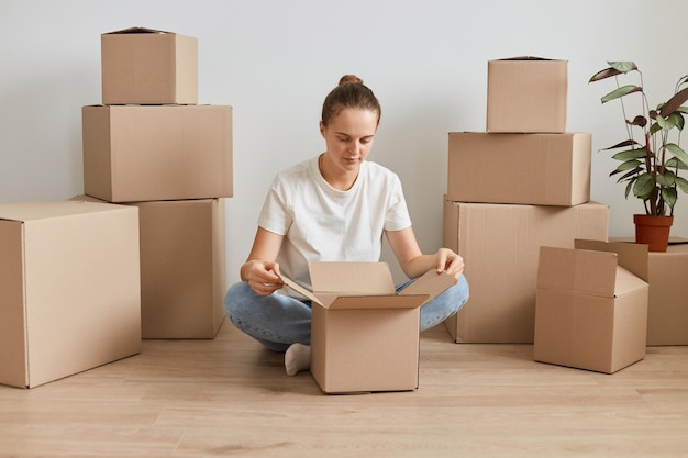 Horizontal shot of Caucasian woman with bun hairstyle wearing white T-shirt sitting on the floor near cardboard boxes with stuff, unpacking packages with personal belonging during relocation.