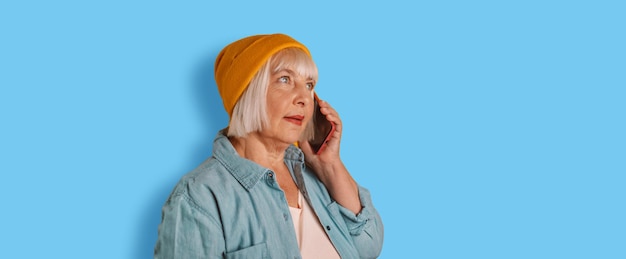 Horizontal shot of cheerful modern senior woman uses phone talking on a cell phone has happy mood over blue color background