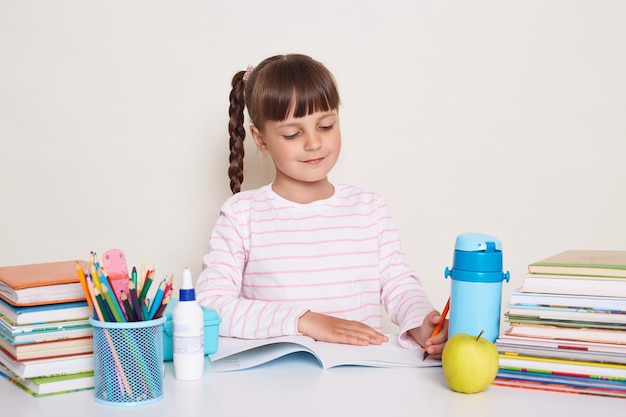 Horizontal shot of cute little schoolgirl with dark hair and braids sitting at table and writing in exercise book posing surrounded with books being concentrated and attentive smiling