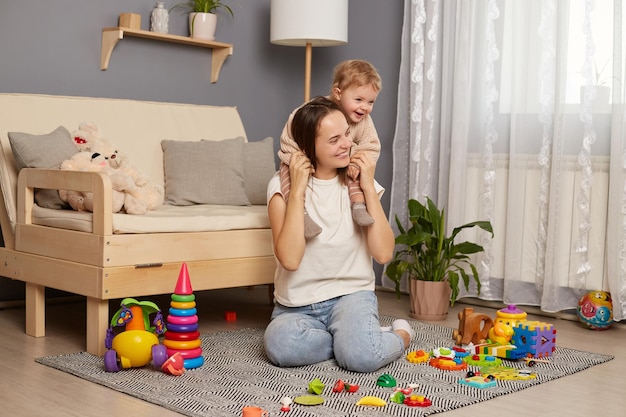 Horizontal shot of dark haired woman wearing white t shirt and jeans holding her toddler baby on her back smiling sitting on carpet on floor cute family play at home